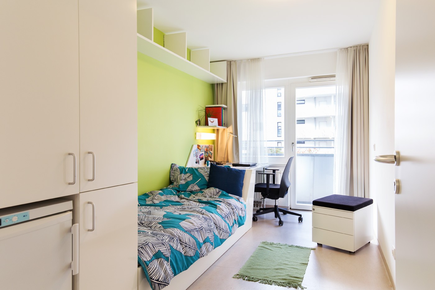 Helles Zimmer mit Einzelbett, Schreibtisch, Schrank, Rollkasten, und Balkonfenster.