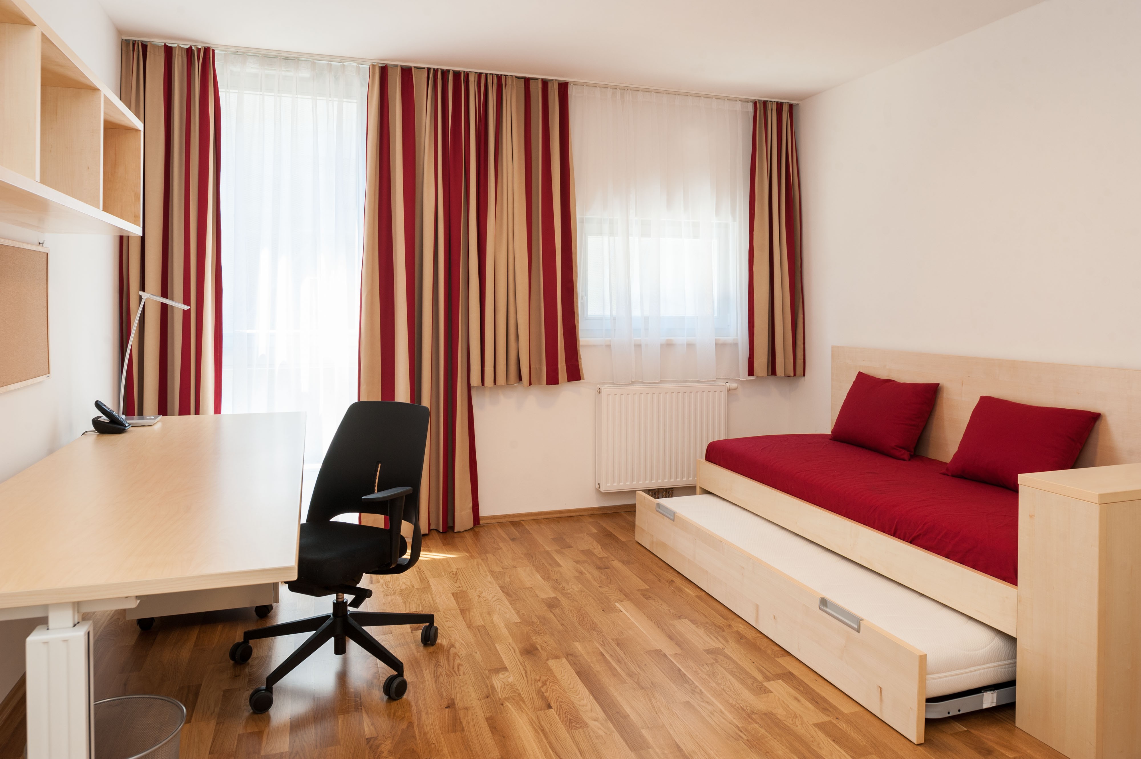 Studentenzimmer mit ausziehbarem Bett, Schreibtisch, Regalen und großen Fenstern