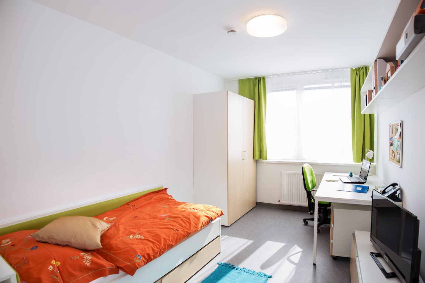 Studentenzimmer mit Einzelbett, Schrank, Schreibtisch mit Stuhl vor Fenster und Fernseher auf niedrigem Regal