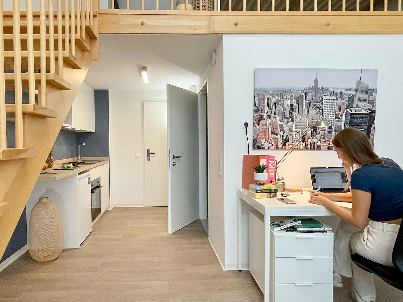 Junge Studentin sitzt an einem Schreibtisch mit Laptop in einem modernen, hellen Studentenheimzimmer mit offener Küche und Holztreppe