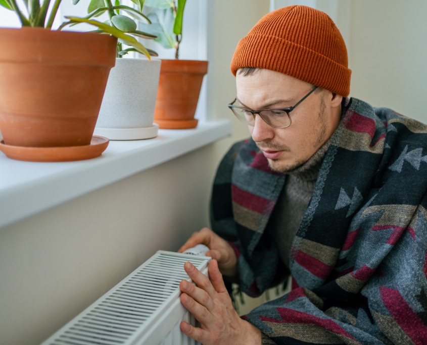 Man suffering from cold at home due to rising energy prices and high heating bills, trying to save money by adjusting the radiator thermostat while wearing warm clothes and a blanket in winter